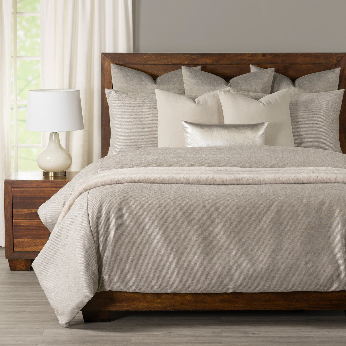 A neatly made bed with light beige and cream bedding, including multiple pillows and a textured throw blanket, set in a bedroom with a dark wood headboard and nightstand.
