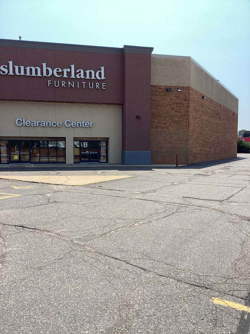 Front exterior of a Slumberland Furniture Clearance Center store with a large cracked asphalt parking lot in the foreground under a clear sky.