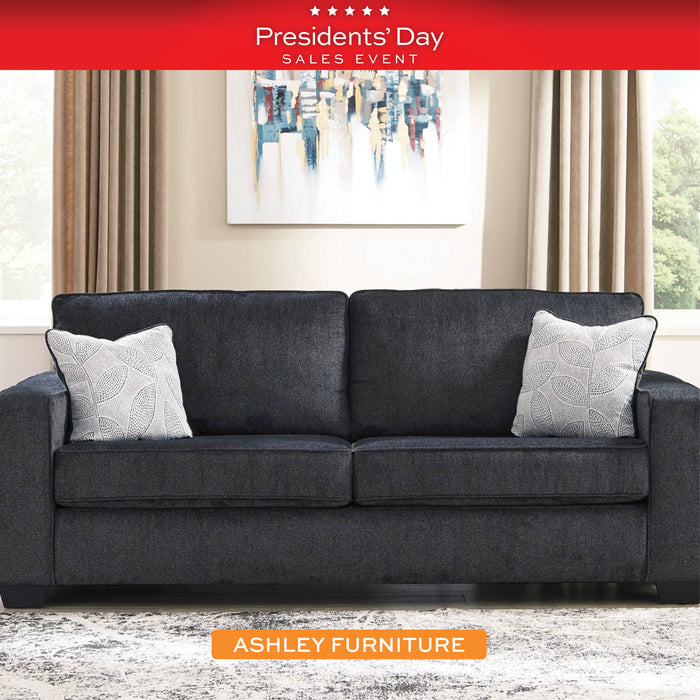 Gray sofa with decorative pillows in a living room setting, featuring Ashley Furniture branding and Presidents' Day sale text.
