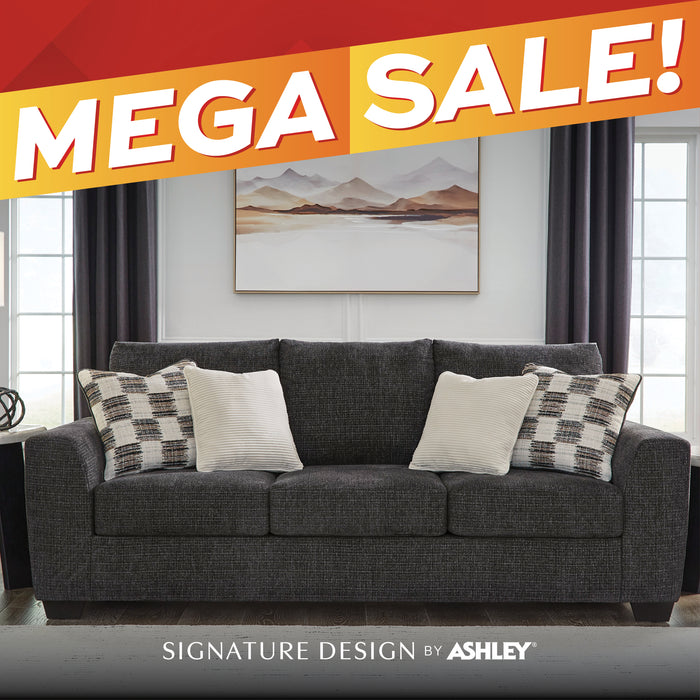 A dark gray upholstered three-seat sofa in a styled living room, with two cream throw pillows and two patterned accent pillows. A large orange and red diagonal banner across the top reads 'MEGA SALE!', and a gray band at the bottom reads 'SIGNATURE DESIGN BY ASHLEY'.