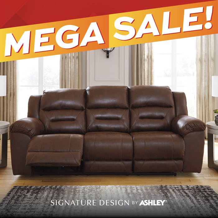 A brown leather reclining three-seat sofa shown in a staged living room with large windows, curtains, side tables, and a rug. A red and orange diagonal banner across the top reads 'MEGA SALE!' and text at the bottom reads 'SIGNATURE DESIGN by ASHLEY'.