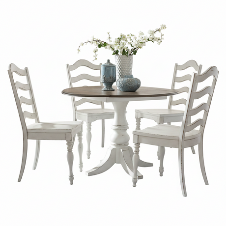 A distressed white dining set featuring a round pedestal table with a dark wood top and four matching ladder-back chairs, styled with a vase of white flowers and two blue-gray ceramic containers on a white background.