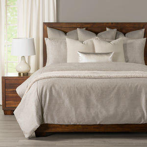 A neatly made bed with light beige and cream bedding, including multiple pillows and a textured throw blanket, set in a bedroom with a dark wood headboard and nightstand.
