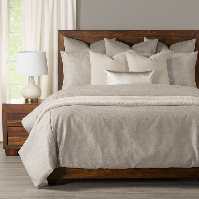 A neatly made bed with light beige and cream bedding, including multiple pillows and a textured throw blanket, set in a bedroom with a dark wood headboard and nightstand.