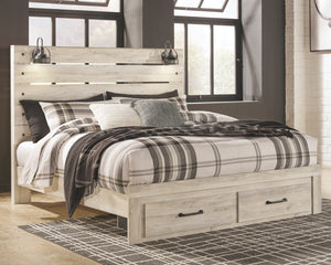 A light whitewashed wood platform bed frame with built-in storage drawers at the foot, styled with plaid and textured bedding in a rustic bedroom setting.