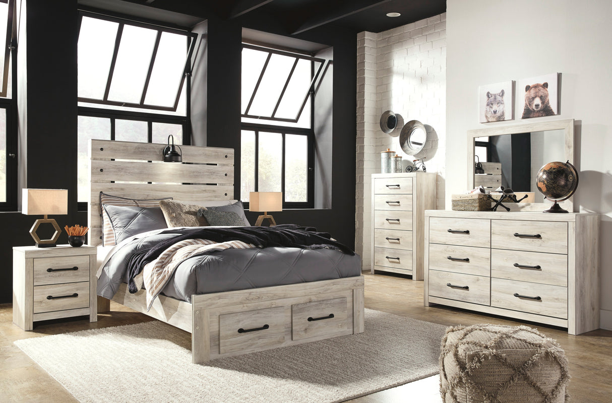A rustic light gray wood bedroom furniture set including a bed with a slatted headboard, two matching nightstands, a dresser, and a tall chest of drawers, styled in a modern loft-style room with black walls and large windows.