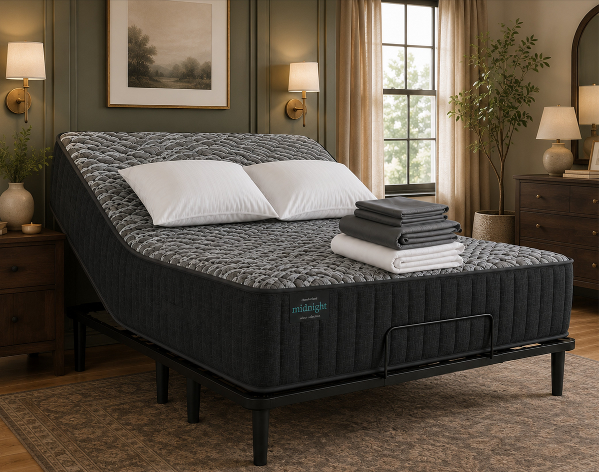 A mattress on a bed frame in a styled bedroom setting, with two white pillows and folded gray and white towels stacked on top. The room has dark wood furniture, lamps, wall art, and a window with curtains.