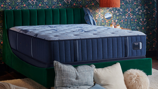 Blue mattress on a green bed frame with decorative pillows against a floral wall.